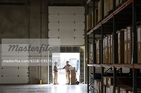 Two warehouse workers shaking hands while standing in the doorway of a loading dock in a large distribution warehouse.