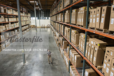 A view from above looking down on a warehouse worker checking inventory of boxes on racks in a distribution warehouse.
