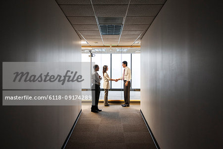 A mixed race group of business people talking and shaking hands in front of a window at the end of a long hallway.