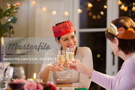 Daughter and senior mother in paper crowns toasting champagne glasses at Christmas dinner table