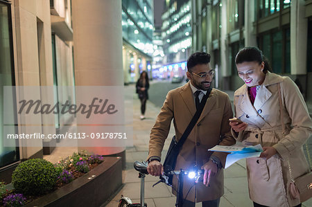 Business people with smart phone and bicycle reviewing paperwork on urban street at night