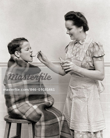 1940s BOY WRAPPED IN BLANKET MOTHER WEARING APRON GIVING HIM A SPOONFUL OF MEDICINE