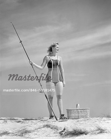 1940s BLOND YOUNG WOMAN WEARING ONE PIECE BATHING SUIT STANDING ON SEASHORE SAND DUNE HOLDING SURF FISHING ROD AND REEL