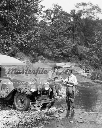 1920s COUPLE FLY FISHING MAN STANDING IN STREAM WOMAN SITTING  ON FENDER OF CAR