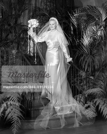 1930s PORTRAIT SMILING BRIDE IN WHITE SATIN WEDDING DRESS AND VEIL WAVING BOUQUET OF FLOWERS STANDING ON STAIRS AMID FERN PLANTS