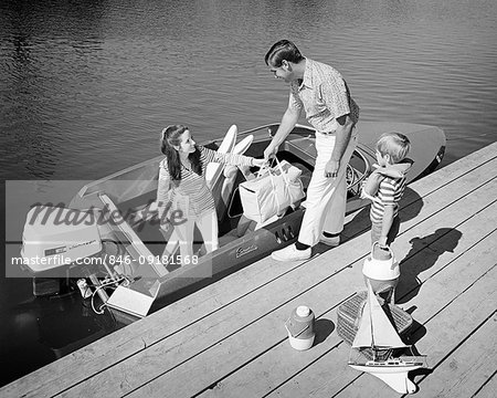 1970s FAMILY OF THREE FATHER MOTHER SON LOADING PICNIC COOLERS TOYS AND WATER SKIS INTO OUTBOARD MOTOR BOAT DOCKSIDE