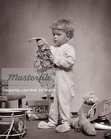 1930s YOUNG BLOND BABY BOY STANDING WEARING WOOL SOCK FOOTED DR. DENTON SLEEPWARE HOLDING A FAVORITE JESTER DOLL AMID OTHER TOYS