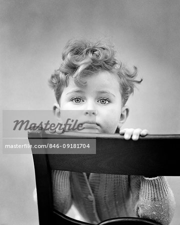 1940s DIFFIDENT CURLY HAIR BOY RESTING CHIN ON BACK OF CHAIR LOOKING AT CAMERA