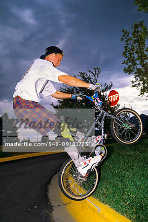 1990s YOUNG MAN WEARING WHITE TEE SHIRT AND SHORTS WITH KNEE PADS JUMPING BICYCLE UP ON CURB