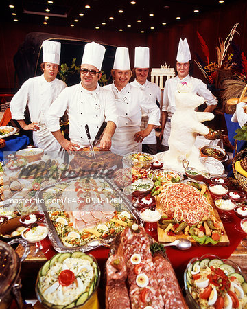 1980s FIVE SMILING CHEFS STANDING BEHIND IMMENSE BUFFET OF MEAT AND MAIN COURSE FOOD PREPARATIONS