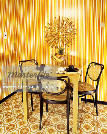 1970s DINING NOOK ORANGE AND YELLOW STRIPED WALLPAPER ORANGE TABLE BROWN WICKER CHAIRS SUNBURST MEDALLION ON WALL
