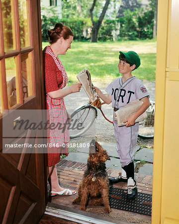 1970s NEWS CARRIER BOY HANDING NEWSPAPER TO WOMAN AT DOORWAY OF HOUSE AIREDALE DOG WATCHING