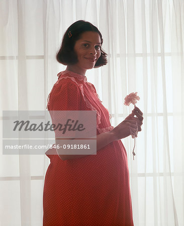 1970s PREGNANT AFRICAN AMERICAN WOMAN LOOKING AT CAMERA SILHOUETTED PROFILE AGAINST SHEER WINDOW LIGHT HOLDING FLOWER SMILING