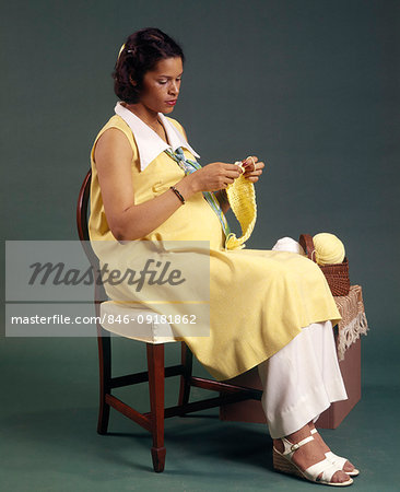 1970s PREGNANT AFRICAN AMERICAN WOMAN SITTING IN STRAIGHT BACK CHAIR KNITTING YELLOW YARN
