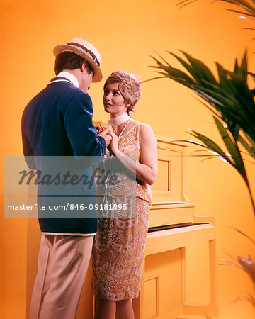 1970s COUPLE MAN WOMAN WEARING 1920s ERA JAZZ AGE CLOTHES HOLDING HANDS STANDING BY PAINTED YELLOW UPRIGHT PIANO