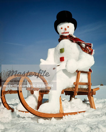 1970s SNOWMAN WEARING TOP HAT SITTING IN EUROPEAN STYLE WOODEN SLED OR SLEIGH LOOKING AT CAMERA