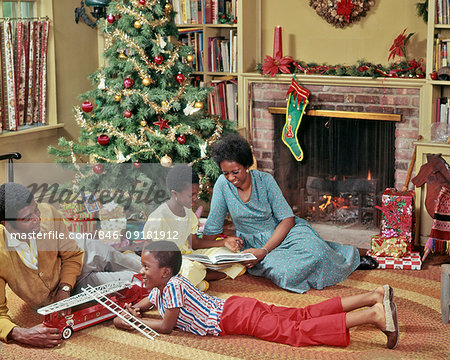 1970s AFRICAN AMERICAN FAMILY OF FOUR ON LIVING ROOM FLOOR PLAYING WITH CHRISTMAS TOYS AND PRESENTS