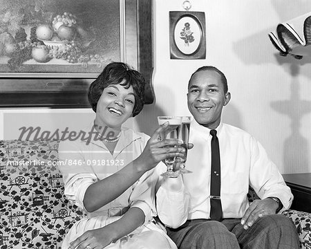 1960s SMILING AFRICAN AMERICAN HUSBAND AND WIFE SITTING IN LIVING ROOM LOOKING AT CAMERA TOASTING WITH RAISED GLASSES OF BEER