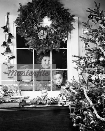 1950s CHILDREN ONE GIRL TWO BOYS LOOKING THROUGH WINDOW AT CHRISTMAS TREE WITH WRAPPED GIFTS WREATH HANGING ABOVE