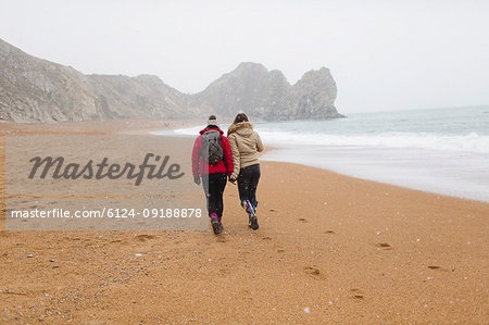 Mother and daughter in warm clothing walking on snowy winter ocean beach