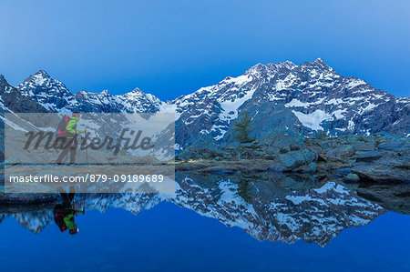 A hiker walks at dusk near a lake that mirrored the mountain range of Mount Disgrazia. Chiareggio valley, Valmalenco, Valtellina, Lombardy, Italy, Europe.