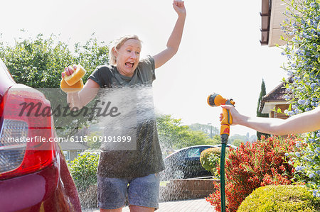 Daughter spraying mother washing car with hose