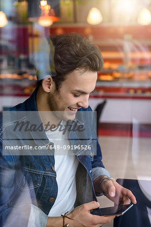 Male student looking at smartphone in cafe window seat, view through window