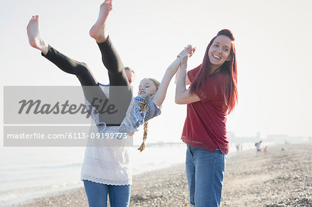 Playful lesbian couple swinging daughter on beach