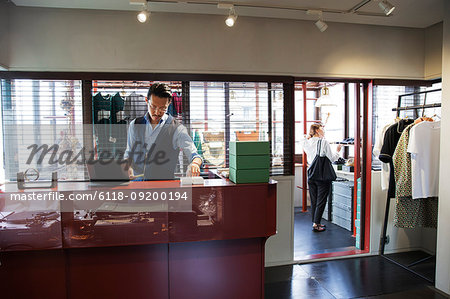 Japanese salesman with moustache wearing glasses standing at counter in clothing store.