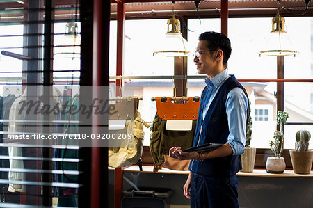 Japanese salesman with moustache wearing glasses standing in clothing store, holding digital tablet.