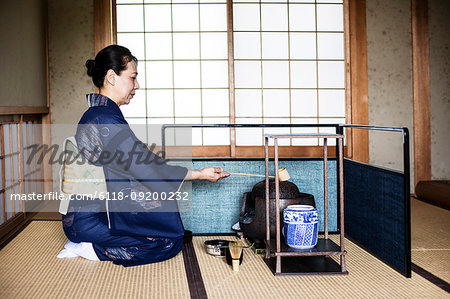 Japanese woman wearing traditional bright blue kimono with cream coloured obi kneeling on floor, using a  Hishaku, a bamboo ladle, during a tea ceremony.