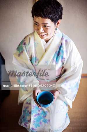 Japanese woman wearing traditional white kimono with blue floral pattern kneeling on floor during tea ceremony, holding blue tea bowl.