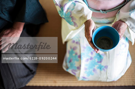 High angle close up of Japanese man and woman wearing traditional white kimono with blue floral pattern kneeling on floor during tea ceremony, holding blue tea bowl.