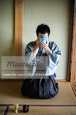 Japanese man wearing traditional kimono kneeling on floor, raising blue tea bowl during tea ceremony.