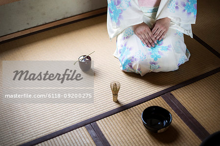 High angle view of Japanese woman wearing traditional white kimono with blue floral pattern kneeling on tatami mat in front of bowl and whisk for tea ceremony.