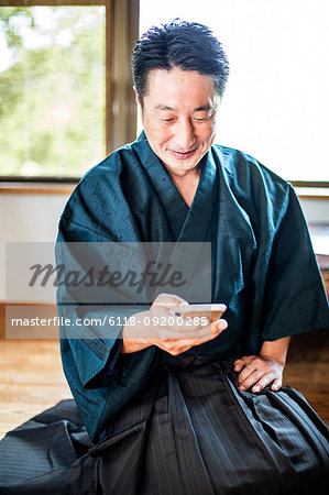 Japanese man wearing kimono sitting on floor in traditional Japanese house, using mobile phone.
