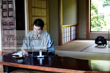 Japanese man wearing kimono sitting on floor in traditional Japanese house, looking at digital tablet.