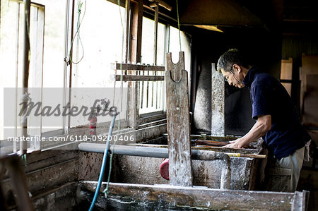 Japanese man in a workshop leaning over a vat of liquid, the traditional Washi papermaking process.