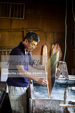 Japanese man in a workshop holding a wooden frame of dried pulp over a basin of water, making traditional Washi paper.