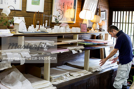 Japanese man in a traditional workshop with a display of handcrafted washi papers and small objects.