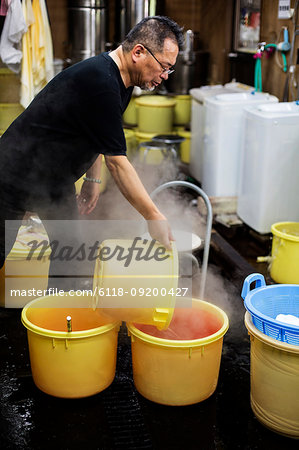 Japanese man standing in a textile plant dye workshop, pouring hot water into yellow plastic buckets.