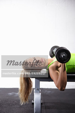 Young female crossfitter lying on bench training with bar bell in gym