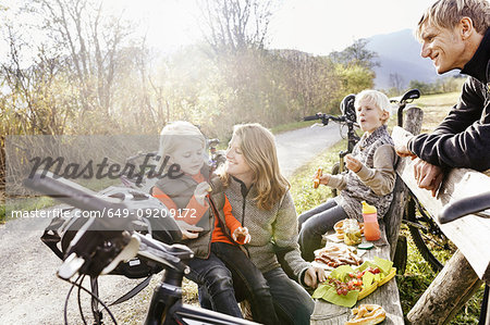 Family with bicycles resting on bench by roadside eating picnic smiling