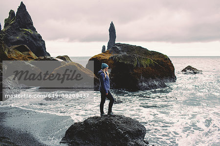 Side view of mid adult woman standing on rock on coastline looking out at ocean, Iceland