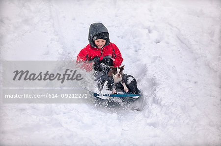 Boy sledging with Boston terrier puppy on snow covered hill smiling