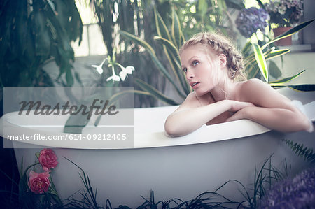 Nude young woman relaxing in bathtub, in bathroom filled with plants