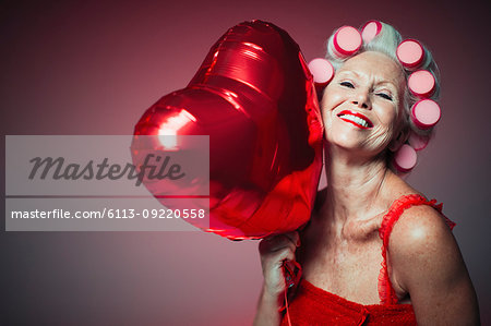 Portrait playful senior woman with hair in curlers holding heart-shape balloon