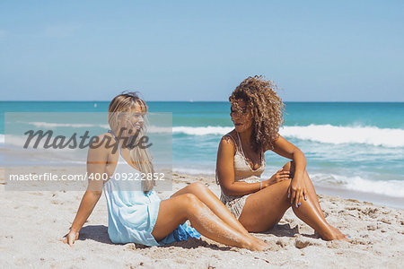 Beautiful happy diverse women sitting on sandy coastline and chatting happily with ocean waves on background.