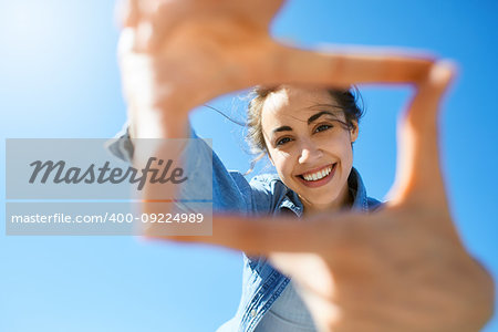 portrait of a young smiling attractive woman in jeans clothes at sunny day on the blue sky background. woman shows a frame from hands like photo. Photo Frame Hands Made By A Hipster young girl