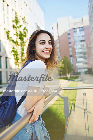 portrait of a young smiling attractive woman in white t-shirt with small city backpack at sunny day on city building background. woman poses in cityscape.